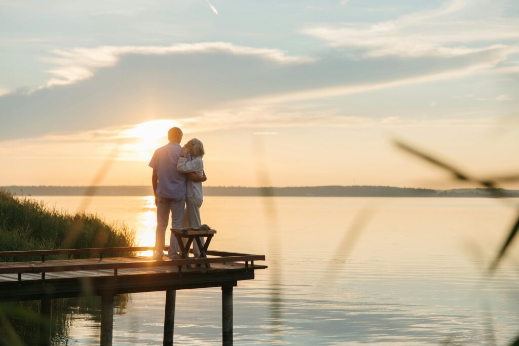 A happily retired couple embracing on a dock as they watch the sunset.